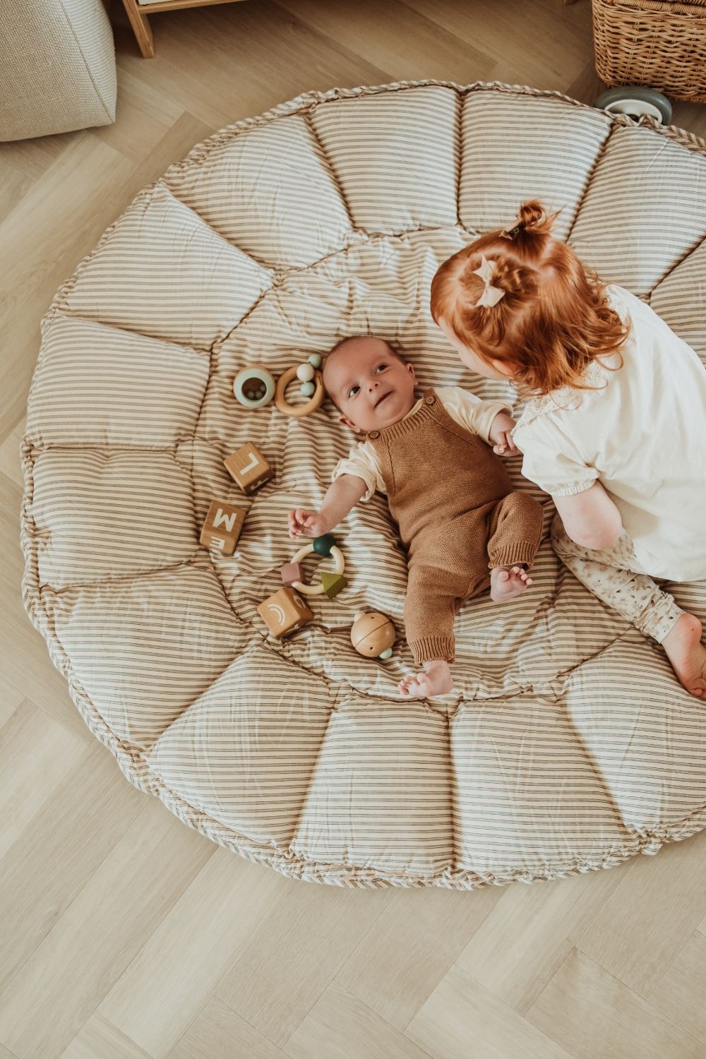bloom stripes blue babymat, open on floor, baby is lying on the mat next to wooden toys, other child is talking to the baby