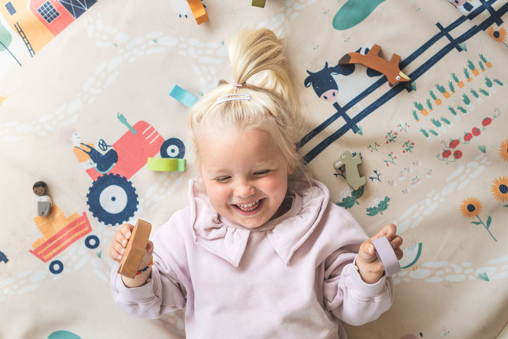 farm storage bag and play mat, child lying on mat, laughing and holding wooden toys in hands